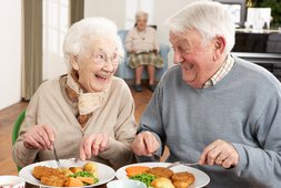 Ein Seniorenpärchen sitzt lachend nebeneinander und genießen Ihr Essen. Es gibt schön grüne Erbsen, Karotten und etwas Paniertes. | © fotolia_monkey_business_30838721